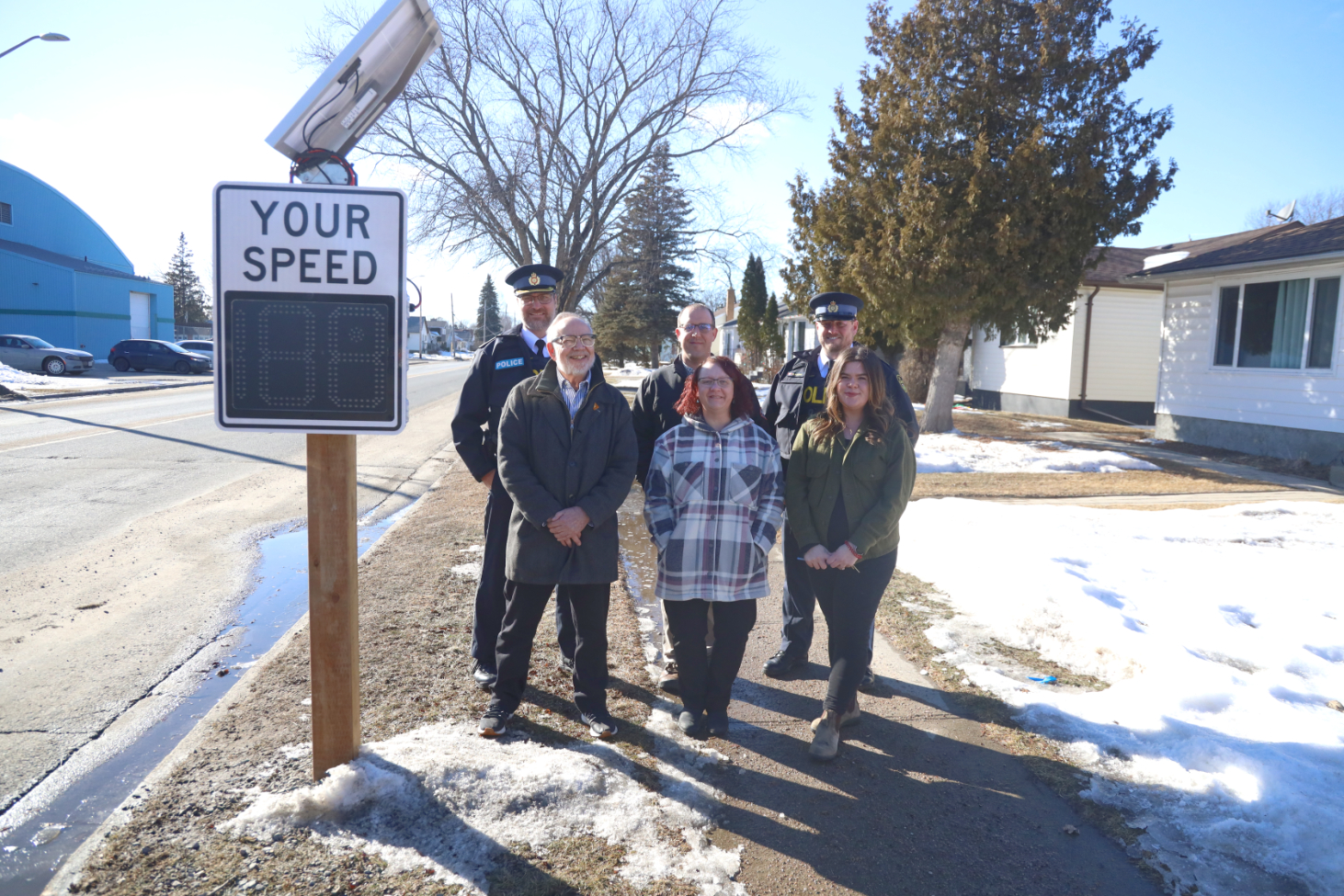 This image shows a group of people, including two uniformed police officers, standing next to a digital speed limit sign displaying "Your Speed" on a suburban street. The sign is equipped with solar panels, indicating it's powered by renewable energy. The group consists of various individuals likely involved in the safety or implementation of this traffic control measure, gathered to either inspect or inaugurate the sign. The environment includes a mix of snowy and clear patches on the ground, suggesting it might be late winter or early spring.